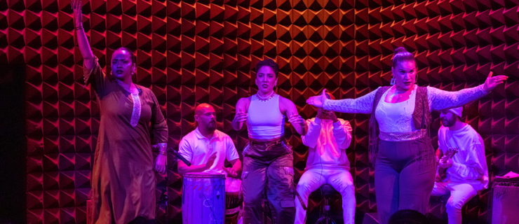 Brinda Guha, Amanda Castro, and Arielle Rosales are lit in pink and purple light as they perform as Soles of Duende at the front of a small stage, two seated percussionists and a guitarist visible behind them. Guha raises an arm to the sky as she looks intensely out at the audience. Castro looks focused as she taps, hands separated but poised to clap. Rosales opens her arms wide in invitation, chin ducked as she looks challengingly out at the audience.