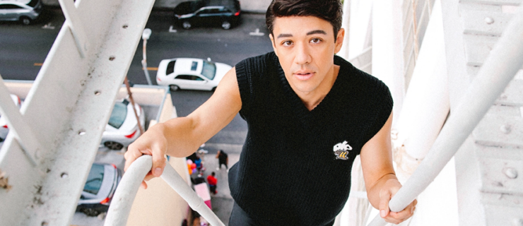 Kyle Hanagami looks curiously up at the camera as he climbs a white-painted fire escape, people and cars visible in the background far below.