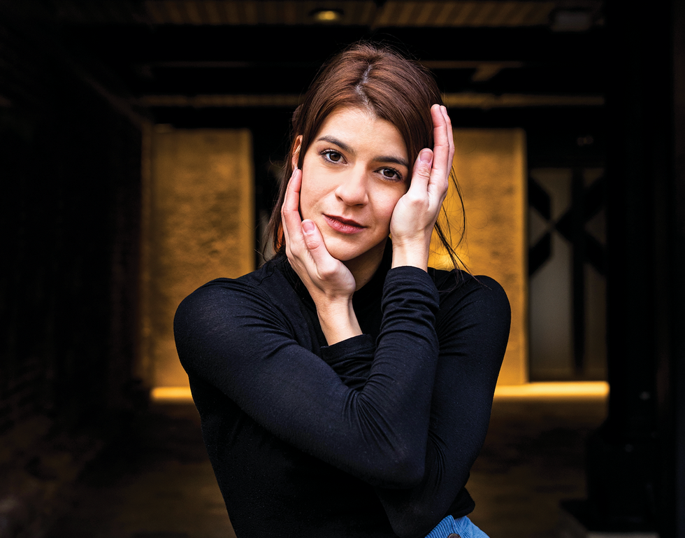 Close-up of Mayara Magri framing her face with her hands. She wears a black turtleneck, hair loosely falling out of an updo.