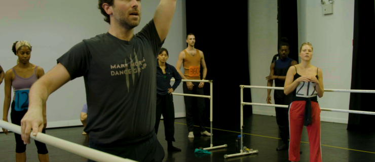 Several dancers with numbers pinned to their rehearsal clothing stand in a dance studio, watching Black—wearing a Mark Morris Dance Group t-shirt—demonstrate a combination. Black's right hand rests on a barre and his left is raised above his head.