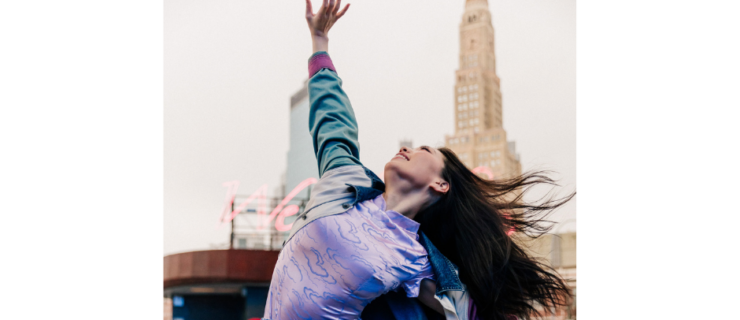 a female dancer wearing a purple top and jeans with a colorful jacket dancing outside on a stone road