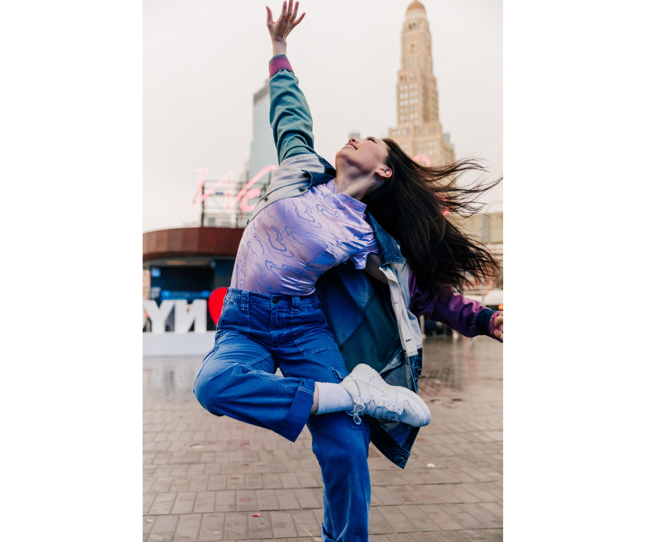 a female dancer wearing a purple top and jeans with a colorful jacket dancing outside on a stone road