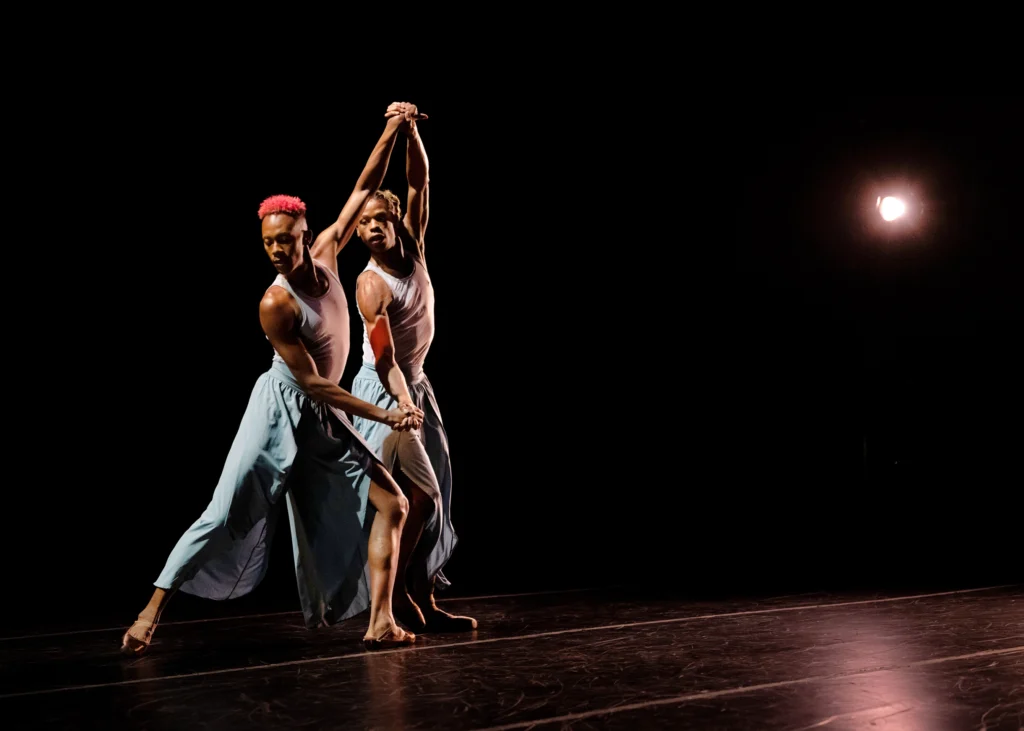 Two dancers in pointe shoes dance side by side, clasping each other's hands. One pair of arms is raised overhead, the other at waist height. The downstage dancer is in fondu tendu back. The other stands with slightly bent knees, as though preparing to move.