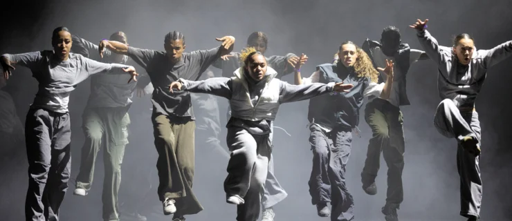 Nine dancers seem to hover over the stage floor, one knee hiking toward their chests, arms extended side, heads and eyes focused forward. The stage is fog-filled; the dancers wear sweats and hoodies in shades of grey and green.