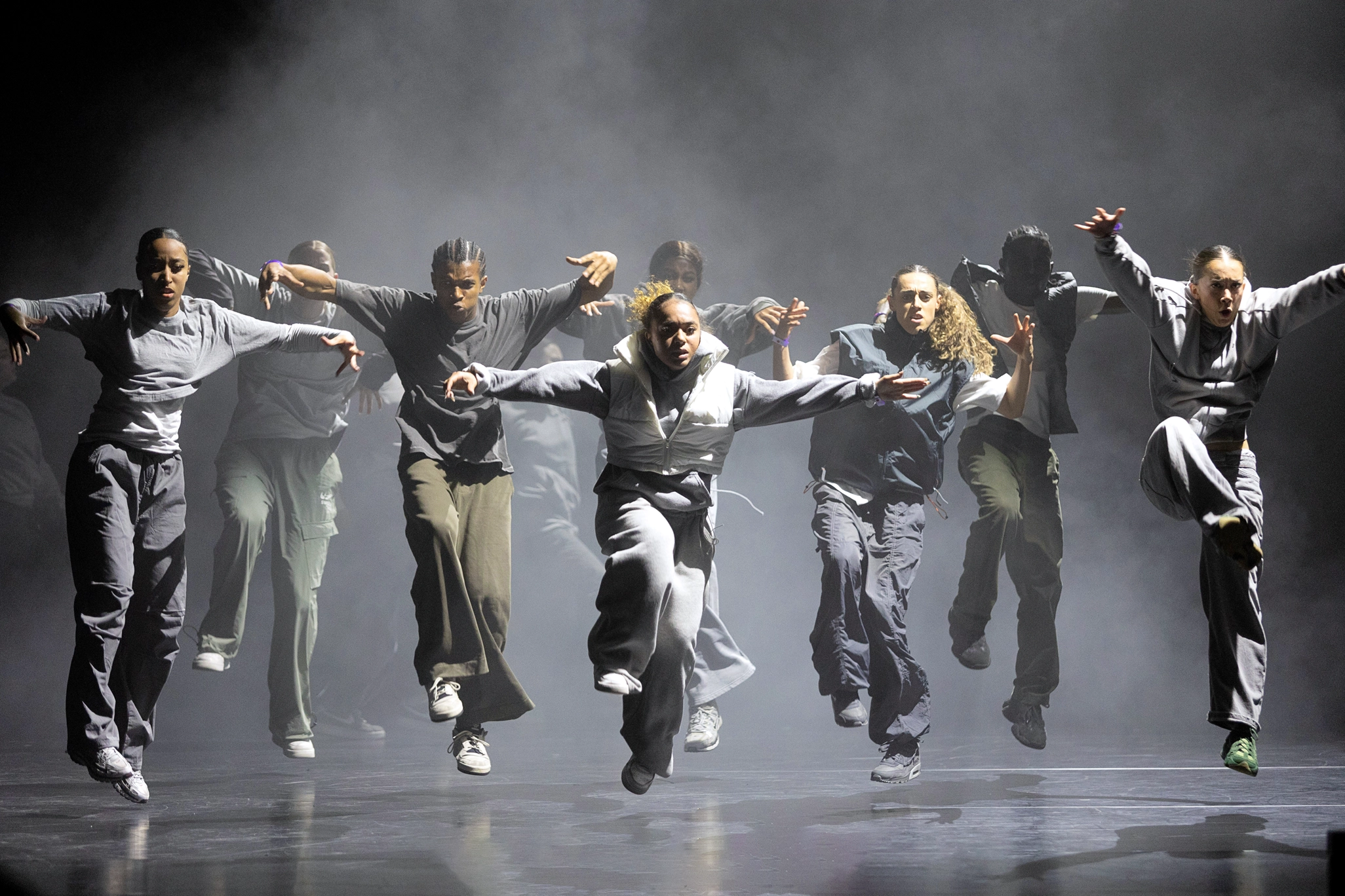 Nine dancers seem to hover over the stage floor, one knee hiking toward their chests, arms extended side, heads and eyes focused forward. The stage is fog-filled; the dancers wear sweats and hoodies in shades of grey and green.