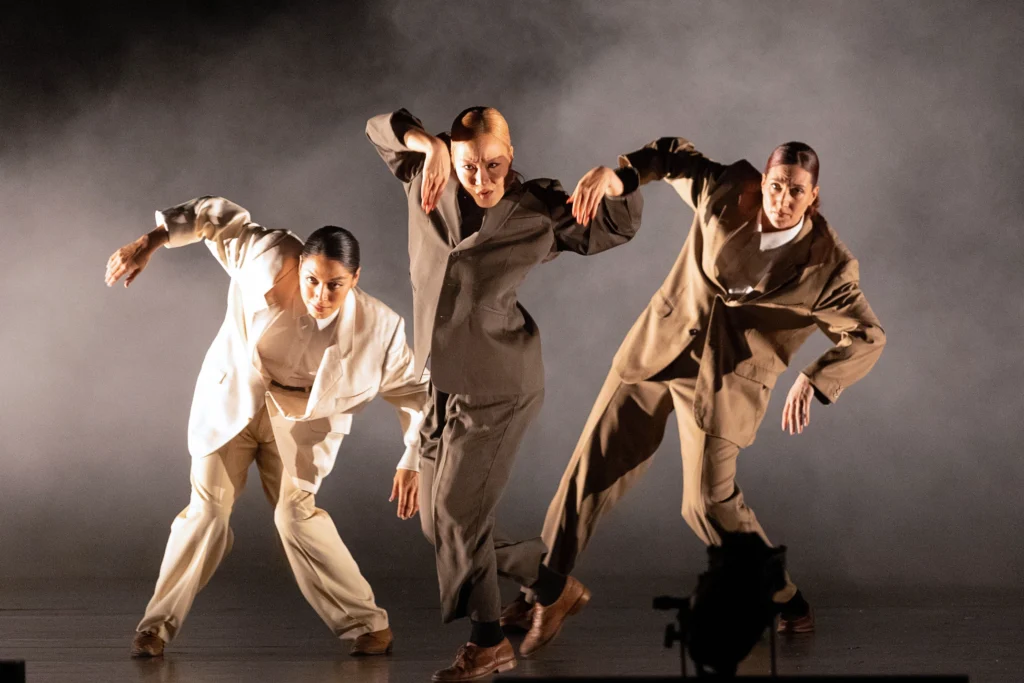 A trio of women in loose-fitting white and brown suits perform on a fog-filled stage. Their knees and elbows bend into angular shapes.