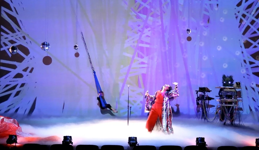 Aretha Aoki dances on a fog-filled stage. She wears red, and her head is tipped back to the ceiling. Ryan MacDonald wears a bear costume with glowing eyes, and stands behind a set of shelves to the right. Another figure sits swinging in a swath of blue fabric suspended from above like a swing.