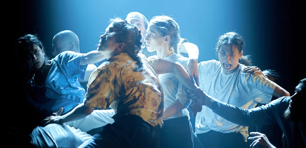 Half a dozen dancers are shown from the waist-up in a tight, chaotic cluster as they are bathed in pale blue light. One appears to be screaming, while others look seriously in a different direction.