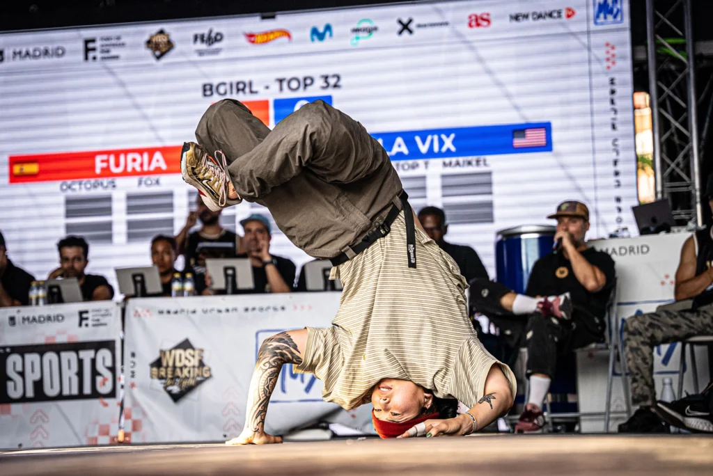 Vicki "La Vix" Chang in competition. Chang freezes on their head, bent arms supporting the balance as their legs bend and lean off-center.