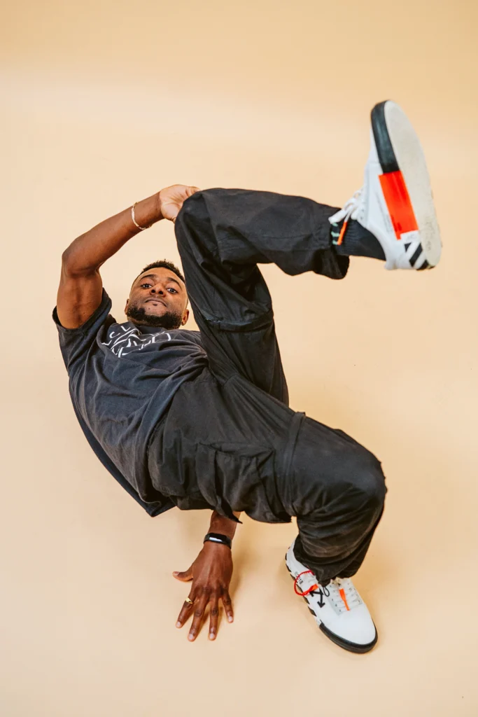 Jeffrey "Jeffro" Louis poses on a nondescript beige background. One foot is planted on the ground while he twists so the opposite side hand can touch the ground next to it. His other arm and leg and bent into the arm to meet. His torso is parallel to the floor; he tips his chin up to look directly at the camera under the frame formed by his arm and thigh.