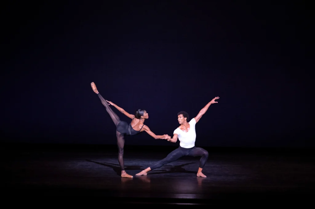 Two Black dancers perform a pas de deux on a dark stage. A woman in pointe shoes tips into a penché on flat, her front arm grasping her partner's, who lunges to the side, away from her.