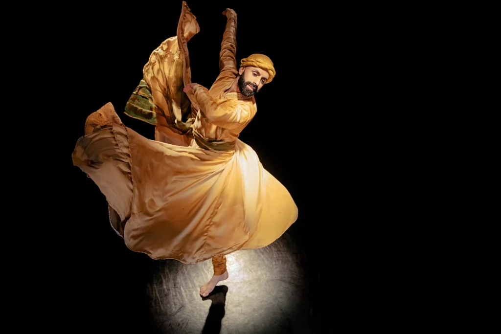 Aakash Odedra dances in a spotlight on a darkened stage, the yellow fabric of his costume flaring around him as he turns on one foot. His expression is serene as he looks down over his shoulder.